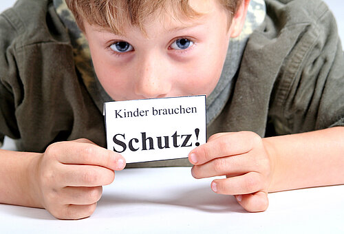 Ein kleiner Junge hält ein kleines Schild mit der Aufschrift Kinder brauchen Schutz! in die Kamera. 