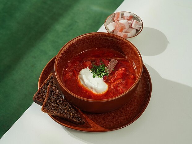 Eine Tonschale vor grün-weißem Hintergrund mit Rote-Beete-Suppe gefüllt. Daneben liegen zwei Scheiben dunkles Brot und dahinter befindet sich eine kleine Schale mit Speckwürfeln.